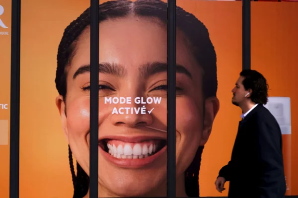 Thomas Ternisien · 07/03/2026 · 2048 x 1362 Cette photographie met en scène une affiche publicitaire géante en arrière-plan, montrant un gros plan souriant avec le texte "MODE GLOW ACTIVE". Le sujet principal est une personne en costume, partiellement visible, marchant devant cette affiche.La composition utilise un cadrage serré sur l'affiche, avec des barres verticales en premier plan. Les couleurs dominantes sont l'orange vif et le blanc. La lumière est vive et contrastée, créant une ambiance dynamique et moderne. Le style est épuré et commercial, évoquant une publicité pour un produit cosmétique ou de soin.