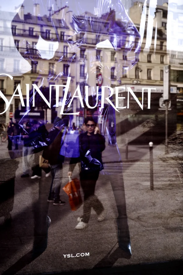 Thomas Ternisien · 07/03/2026 · 2048 x 3080 Cette photographie montre une vitrine reflétant des passants devant un magasin Saint Laurent. Le sujet principal est le reflet des personnes marchant, avec leurs silhouettes floues et dynamiques. La composition utilise un cadrage serré sur la vitrine, capturant une partie de la rue parisienne.La lumière est tamisée, avec des teintes douces et des ombres contrastées. Les couleurs dominantes sont les tons froids (bleus, gris) et quelques éclats chauds (orange, rouge). Le style est urbain et moderne, évoquant une ambiance chic et animée typique des grandes villes.