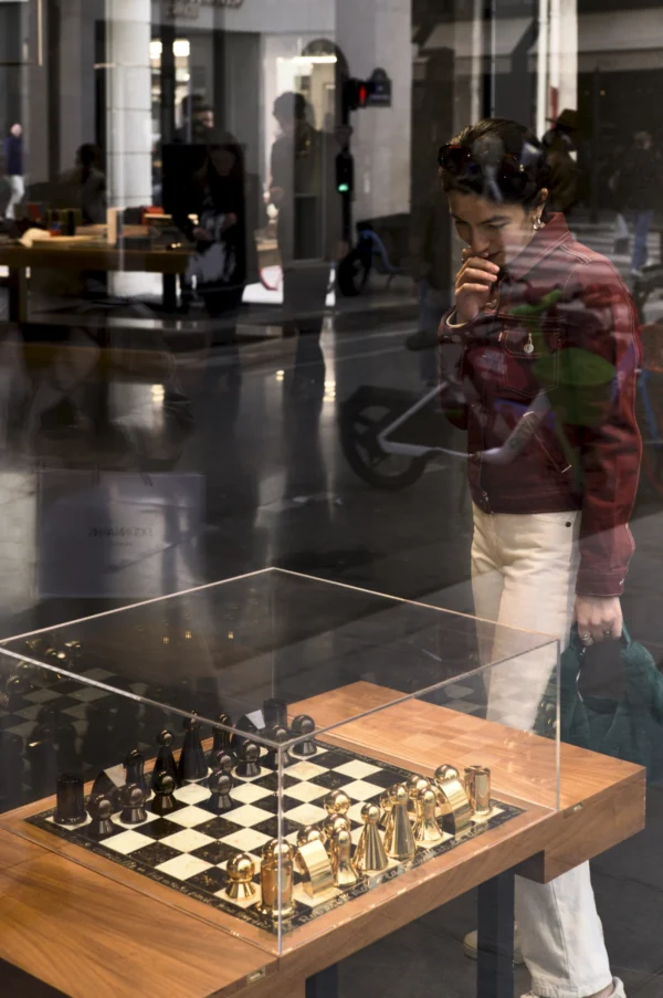 Thomas Ternisien · 07/03/2026 · 2048 x 3082 Cette photographie met en scène un **jeu d'échecs géant** sous une vitrine, comme sujet principal. La composition montre une personne en arrière-plan, partiellement visible, qui semble observer le jeu.Le cadrage est serré sur la table et le jeu, avec une profondeur de champ qui floute légèrement l'arrière-plan. La lumière est douce et naturelle, éclairant uniformément la scène. Les couleurs dominantes sont neutres (blanc, noir, bois) avec des touches dorées sur les pièces.L’ambiance est calme et artistique, évoquant un espace muséal ou une exposition. Le style est épuré et contemporain.