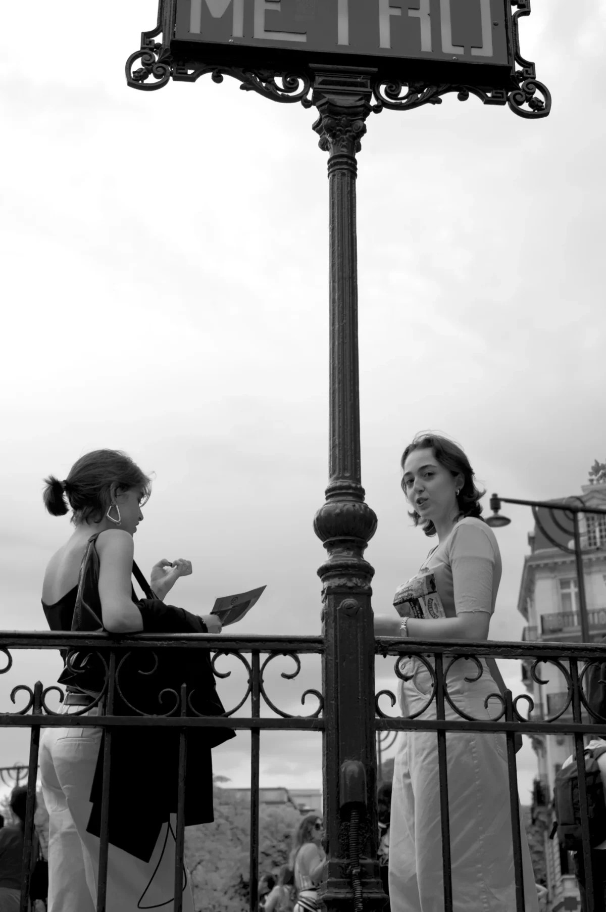 Thomas · 07/09/2025 · 2724 x 4096 Cette photographie en noir et blanc montre deux femmes comme sujets principaux, assises sur une balustrade. La composition est symétrique, avec un lampadaire central portant un panneau "Métro".Le cadrage est serré sur elles, avec un arrière-plan flou. La lumière est douce, probablement naturelle, créant des contrastes modérés. Les couleurs dominantes, bien que monochromes, suggèrent des tons clairs et sombres typiques du noir et blanc.Le style évoque une ambiance vintage, avec une touche de nostalgie. L'atmosphère est calme et intime, renforcée par le cadre urbain et la pose détendue des femmes.