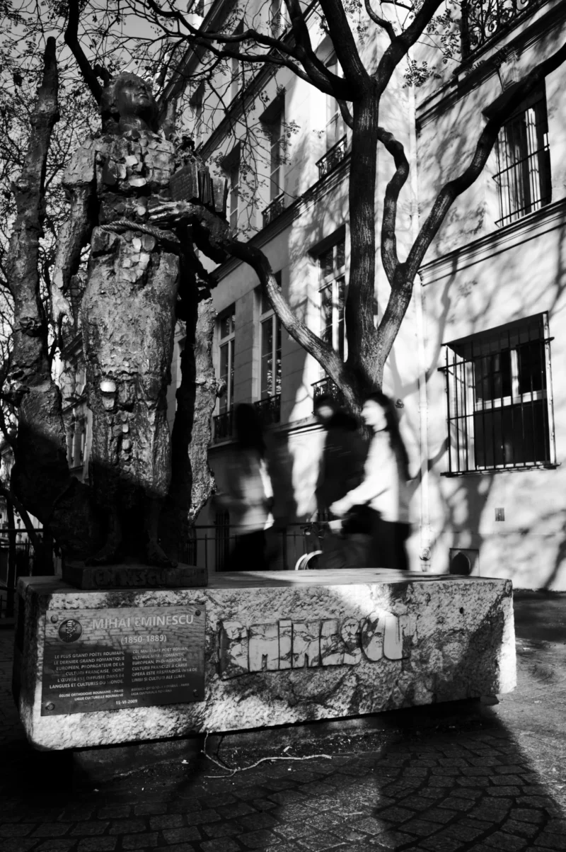 Thomas · 21/03/2026 · 2724 x 4096 Cette photographie en noir et blanc capture une scène urbaine mystérieuse. Le sujet principal semble être une silhouette floue d’une personne, partiellement cachée par les branches d’un arbre et les murs. La composition joue sur les contrastes entre ombre et lumière, accentuant une ambiance dramatique. Le cadrage inclut des éléments architecturaux, comme une façade de bâtiment et des escaliers en pierre couverts de graffitis.La lumière, tamisée et contrastée, crée des jeux d’ombres intrigants. Les couleurs dominantes, bien que limitées au noir et blanc, renforcent les textures et les contrastes. Le style rappelle le photojournalisme ou une approche artistique documentaire, évoquant une ambiance mélancolique et onirique.