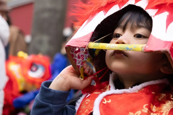 Laurence · 1350 x 900 Cette photographie met en scène une jeune enfant, sujet principal, vêtue d’un habit traditionnel rouge orné de motifs dorés, typique des célébrations asiatiques comme le Nouvel An chinois. Elle tient un lampion rouge et mange un encas.Le cadrage est serré, centré sur son visage, avec un arrière-plan flou où l’on devine d’autres personnes et décorations festives. La lumière est douce et naturelle, mettant en valeur les couleurs vives. Les teintes dominantes sont le rouge éclatant et le doré, renforçant l’ambiance festive et chaleureuse. Le style est documentaire et spontané, capturant un moment authentique de célébration.