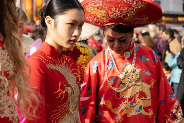 Laurence · 1350 x 900 Cette photographie met en scène deux personnes vêtues de costumes traditionnels asiatiques, probablement vietnamiens. Le sujet principal est une femme en robe rouge brodée, accompagnée d’un homme portant un chapeau ornés et une veste richement décorée de motifs dorés.La composition est serrée, centrée sur eux, avec une ambiance intimiste malgré la foule en arrière-plan. Le cadrage est rapproché, mettant en valeur les détails des tenues. La lumière est douce, naturelle, éclairant les couleurs vives. Les couleurs dominantes sont le rouge éclatant et l’or, typiques des célébrations traditionnelles. L’ambiance est festive et culturelle, évoquant une cérémonie ou une fête.