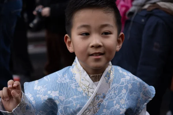Laurence · 1350 x 900 Cette photographie met en scène un jeune garçon comme sujet principal, vêtu d’un costume traditionnel bleu pâle à motifs floraux, évoquant une tenue culturelle asiatique. Le cadrage est serré sur son visage et sa main, isolant le sujet des autres personnes floues en arrière-plan.La lumière semble naturelle et douce, mettant en valeur les détails du costume. Les couleurs dominantes sont les bleus pastel et les blancs, avec des touches de rouge et noir en arrière-plan. L’ambiance est chaleureuse et solennelle, suggérant une cérémonie ou un événement traditionnel. Le style est documentaire et captif, avec une attention portée aux expressions et détails vestimentaires.