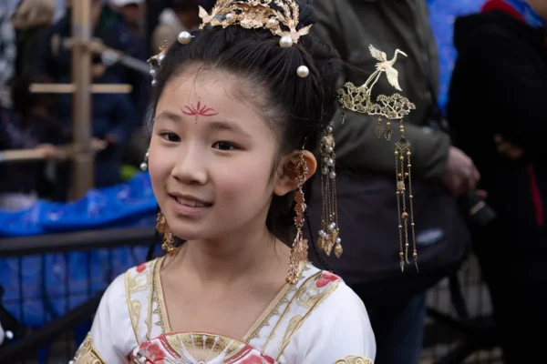 Laurence · 1350 x 900 Cette photographie met en scène une jeune fille comme sujet principal, vêtue d’un costume traditionnel orné et élégant. Elle porte une coiffe richement décorée et des bijoux dorés.La composition est centrée sur son visage souriant, avec un cadrage serré. L’arrière-plan est flou, mettant en valeur la jeune fille. La lumière est douce, éclairant son visage de manière naturelle. Les couleurs dominantes sont les tons chauds (or, rouge, blanc) et les teintes pastel.Le style évoque une ambiance culturelle et festive, probablement liée à une célébration traditionnelle. L’atmosphère est joyeuse et chaleureuse.