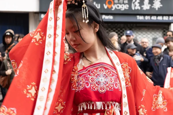 Laurence · 1350 x 900 Cette photographie met en scène une femme vêtue d’un costume traditionnel japonais, probablement un kimono de cérémonie, avec une robe rouge ornée de motifs blancs et une longue écharpe. Elle semble absorbée ou en mouvement, le visage partiellement caché.La composition est centrée sur elle, avec un arrière-plan flou montrant une foule et des enseignes. Le cadrage est serré, mettant l’accent sur les détails de sa tenue. La lumière est naturelle, douce, et les couleurs dominantes sont le rouge vif et le blanc, évoquant une ambiance à la fois traditionnelle et festive. L’ensemble dégage une impression de solennité et de culture.