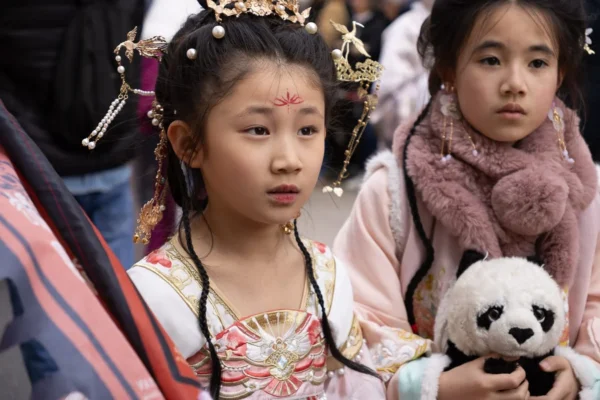 Laurence · 1350 x 900 Cette photographie met en scène deux jeunes filles, dont l’une est le sujet principal. Elle porte un costume traditionnel richement ornné, avec des motifs colorés et des accessoires dorés, tenant un ours en peluche blanc. La composition est serrée, centrée sur les visages et les détails vestimentaires. Le cadrage est rapproché, créant une ambiance intime.La lumière semble naturelle, douce et légèrement tamisée. Les couleurs dominantes sont les tons chauds (or, rouge) et pastel (rose, blanc), évoquant une touche culturelle et festive. Le style est documentaire, capturant un moment authentique et traditionnel. L’ambiance est à la fois joyeuse et solennelle.