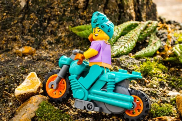 Laurence Givry · 14/03/2026 · 1350 x 900 Cette photographie met en scène une figurine Lego représentant un personnage vêtu d’un costume coloré, coiffé d’un chapeau vert, assis sur un scooter turquoise. La composition est simple et centrée, avec un cadrage serré sur le sujet. L’ambiance est naturelle, avec un arrière-plan de rochers et de mousse.Les couleurs dominantes sont vives : turquoise, vert, orange et violet. La lumière semble naturelle, douce et légèrement tamisée, créant des ombres douces. Le style est ludique et créatif, évoquant une touche fantastique et poétique.