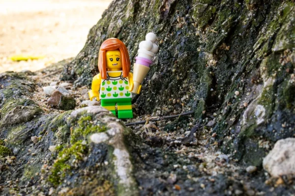 Laurence Givry · 14/03/2026 · 1350 x 900 Cette photographie met en scène une figurine Lego représentant une personne aux cheveux roux, vêtue d’une robe à motifs. Le sujet principal est assis sur une souche moussue, entourée de rochers et de végétation.Le cadrage est serré, centré sur la figurine, avec un arrière-plan naturel flou. La lumière semble naturelle, douce et légèrement tamisée, créant des ombres douces. Les couleurs dominantes sont les verts (mousse, végétation) et les teintes vives de la figurine (vert, jaune, rouge).Le style est minimaliste et photographique, évoquant une ambiance paisible et presque onirique, mêlant nature et jouet.