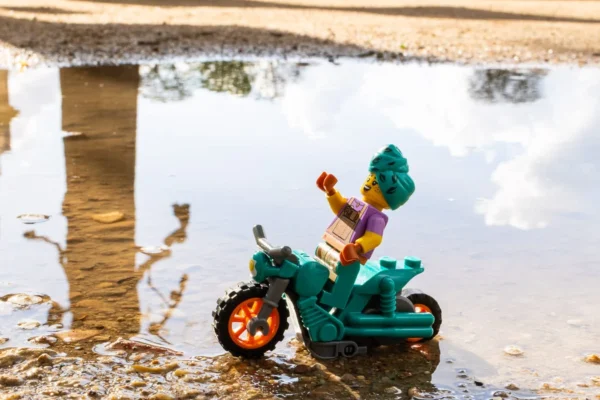 Laurence Givry · 14/03/2026 · 1350 x 900 Cette photographie met en scène un petit personnage Lego assis sur une moto miniature. Le sujet principal est ce jouet, reflété dans une flaque d’eau, créant un effet miroir.La composition joue sur la symétrie et la réflexion, renforcée par le cadrage serré. La lumière naturelle semble douce, éclairant les détails du jouet et de son reflet. Les couleurs dominantes sont vives : vert, bleu, orange et jaune, typiques des Lego.Le style est simple et ludique, évoquant une ambiance poétique et presque onirique grâce au reflet. L’image dégage une touche de nostalgie et de magie du quotidien.