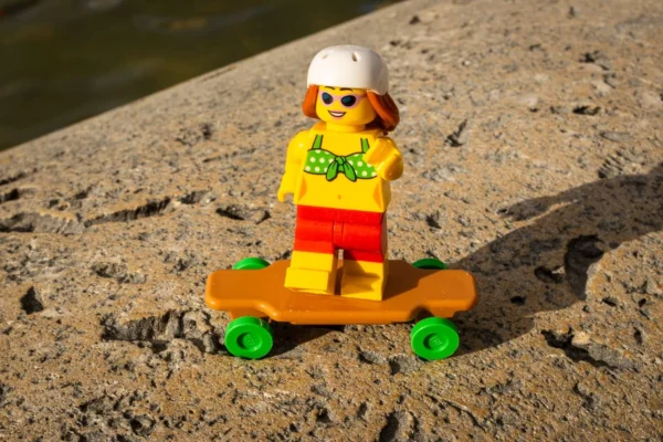 Laurence Givry · 14/03/2026 · 1350 x 900 Cette photographie montre une figurine Lego représentant un surfeur, vêtu d’un maillot jaune et de shorts rouges, sur une planche à roulettes orange avec des roues vertes. Le sujet est positionné sur une surface sableuse près de l’eau.La composition est simple et centrée, avec un cadrage serré mettant en valeur la figurine. La lumière naturelle, douce et légèrement rasante, crée des ombres marquées. Les couleurs dominantes sont vives : jaune, orange, rouge et vert, typiques des Lego. Le style est ludique et minimaliste, évoquant une ambiance détendue et estivale.
