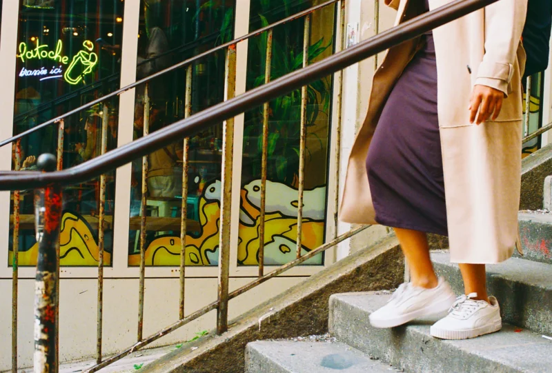 Nadia · 3601 x 2433 Cette photographie met en scène une personne vue de dos, vêtue d’une robe longue et de baskets blanches, montant des escaliers métalliques. Le cadrage est serré sur ses jambes et le garde-corps, créant une composition dynamique.Les couleurs dominantes sont des tons pastel (violet, jaune) et des teintes neutres. La lumière semble naturelle, douce et légèrement tamisée, évoquant une ambiance urbaine détendue. Les graffitis colorés en arrière-plan ajoutent une touche artistique et vivante. Le style est à la fois spontané et esthétique, avec une ambiance moderne et décontractée.