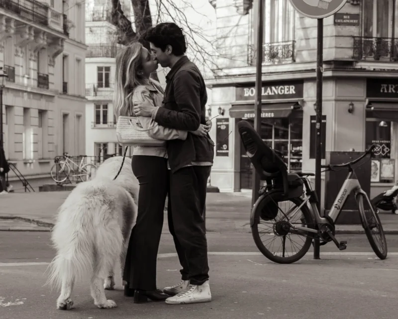 Alice · 21/03/2026 · 2228 x 1782 Cette photographie met en scène un couple en train de s'embrasser tendrement, entouré d'un chien blanc. Le sujet principal est cette interaction émotionnelle.La composition est simple et naturelle, avec un cadrage serré sur les protagonistes. L'arrière-plan montre une rue urbaine avec des vélos et des bâtiments, ajoutant une touche de vie quotidienne.La lumière est douce, probablement naturelle, créant des contrastes modérés. Les couleurs dominantes sont en noir et blanc, ce qui accentue les textures et les émotions.Le style est documentaire et intimiste, évoquant une ambiance chaleureuse et romantique.