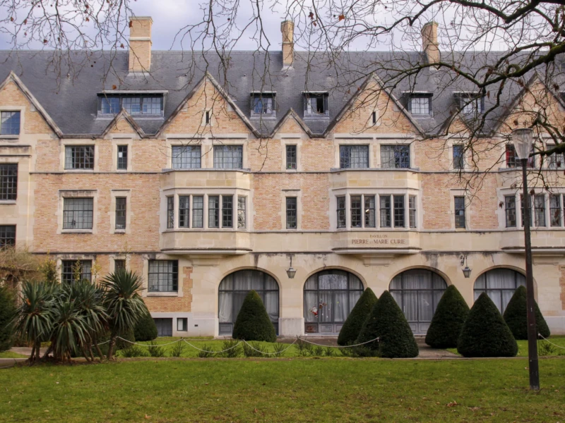 Cité universitaire Cette photographie montre un bâtiment de style architectural classique, probablement une résidence ou un hôtel particulier. Le sujet principal est l’imposante façade en pierre claire, avec ses fenêtres à petits carreaux et ses arcs en plein cintre au rez-de-chaussée.La composition est symétrique, avec un cadrage centré sur le bâtiment. La lumière est naturelle, douce et légèrement voilée, créant des ombres discrètes. Les couleurs dominantes sont les tons beiges et gris de la pierre, contrastant avec le vert du gazon et les touches de brun des arbres.L’ambiance est élégante et sereine, évoquant le calme et le raffinement.