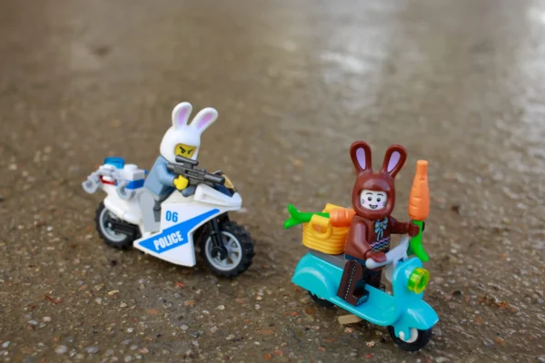 Cédric Manuel · 14/03/2026 · 2048 x 1365 Cette photographie montre deux figurines en plastique représentant des lapins. Le sujet principal est un lapin policier sur une moto et un autre lapin en tenue de travail sur un scooter turquoise.La composition est symétrique et dynamique, avec un cadrage serré. La lumière naturelle est douce, créant des ombres légères. Les couleurs dominantes sont vives : bleu, turquoise, blanc et orange.Le style est ludique et enfantin, évoquant un univers de jouets. L’ambiance est joyeuse et fantaisiste, rappelant un jeu ou une scène de fiction.