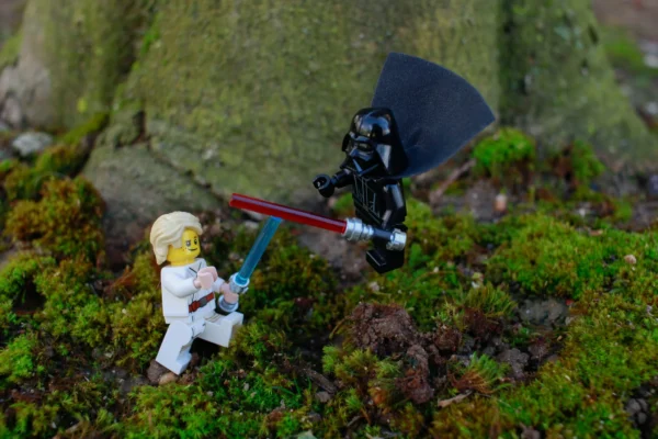 Cédric Manuel · 14/03/2026 · 2048 x 1365 Cette photographie montre deux figurines Lego représentant des personnages de *Star Wars* : un personnage en tenue blanche (probablement Luke Skywalker) et un stormtrooper noir. Le sujet principal est leur confrontation, avec le stormtrooper brandissant une hache et Luke tenant un sabre laser.La composition est dynamique, avec un cadrage serré sur les figurines posées sur un sol moussus et rocheux. La lumière naturelle met en valeur les détails, créant des ombres douces. Les couleurs dominantes sont les verts (mousse), gris (roche), et les teintes vives des tenues des personnages. Le style est ludique et miniature, évoquant une ambiance épique et fantastique.
