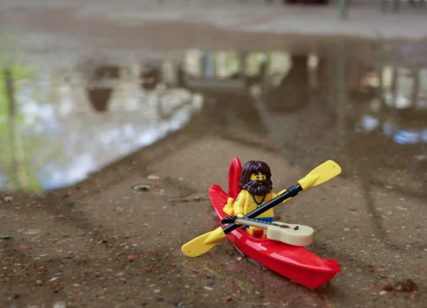 Cédric Manuel · 14/03/2026 · 2048 x 1478 Cette photographie met en scène un petit avion en plastique miniature, sujet principal, posé sur une surface terreuse près d’une flaque d’eau reflétant son image. La composition est simple et centrée, avec un cadrage serré qui isole l’avion.La lumière semble naturelle, douce et légèrement tamisée, créant des ombres discrètes. Les couleurs dominantes sont vives : rouge, jaune et noir pour l’avion, contrastant avec les tons neutres du sol et de l’eau. Le style est épuré et minimaliste, évoquant une ambiance ludique et poétique, presque onirique.
