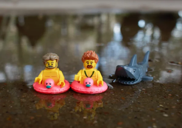Cédric Manuel · 14/03/2026 · 2048 x 1451 Cette photographie montre trois petites figurines en plastique posées sur une surface réfléchissante et humide.Le sujet principal est deux figurines humanoïdes vêtues de combinaisons orange, flottant sur des cerceaux roses, accompagnées d’un requin bleu. La composition est simple et symétrique, avec un cadrage serré. La lumière naturelle est douce, créant des reflets sur l’eau.Les couleurs dominantes sont le rose, l’orange et le bleu. Le style est minimaliste et ludique, évoquant une ambiance poétique et presque onirique, comme une scène de jeu ou de baignade miniature.