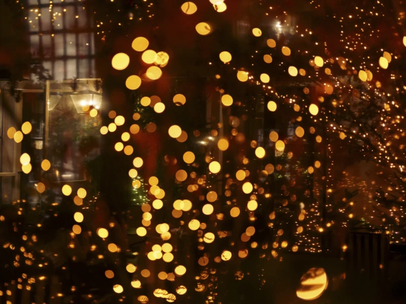 Une nuit dans le Marais Cette photographie capture une ambiance festive nocturne. Le sujet principal semble être une multitude de **lumières dorées et scintillantes**, évoquant des guirlandes ou des feux d'artifice. La **composition** est floue et abstraite, avec un **cadrage serré** qui isole les points lumineux.Les **couleurs dominantes** sont des tons chauds (jaunes, ors), créant une ambiance chaleureuse et magique. En arrière-plan, des silhouettes floues suggèrent une architecture urbaine. Le **style** est artistique et poétique, tandis que l’**ambiance** est onirique et festive, presque hypnotique.