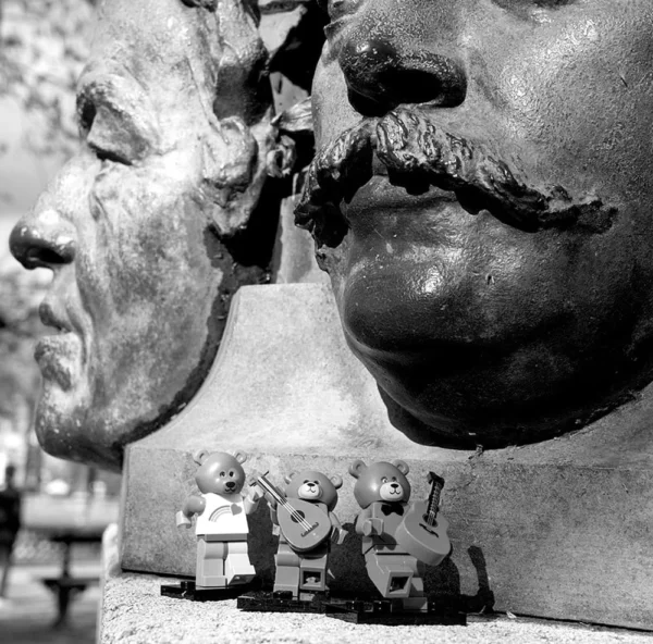 Pascale CHESNOT · 18/03/2026 · 1000 x 987 Cette photographie montre une sculpture monumentale en gros plan, représentant un visage moustachu et sévère. En bas, une petite scène intégrée figure trois personnages jouant de la musique, créant un contraste saisissant.La composition joue sur les échelles et les contrastes. Le cadrage serré met en valeur les détails du visage, tandis que la lumière, naturelle et contrastée, accentue les textures. En noir et blanc, les tons varient du clair au foncé, renforçant l’ambiance dramatique et mystérieuse. Le style semble artistique et provocateur, mêlant grandeur et ironie.