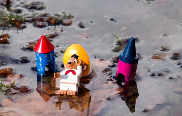 Pascale CHESNOT · 14/03/2026 · 1000 x 636 Cette photographie montre une scène miniature avec des figurines Lego sur un fond évoquant une surface liquide, probablement de la résine ou de l’eau.Le sujet principal est une figurine Lego habillée en marin, entourée de trois petits bonnets colorés (rouge, jaune et bleu). La composition est symétrique et centrée, avec un cadrage serré sur les figurines. La lumière semble naturelle et douce, créant des ombres légères. Les couleurs dominantes sont vives et contrastées : rouge, bleu, jaune et violet.Le style est ludique et créatif, évoquant une ambiance fantastique et poétique.