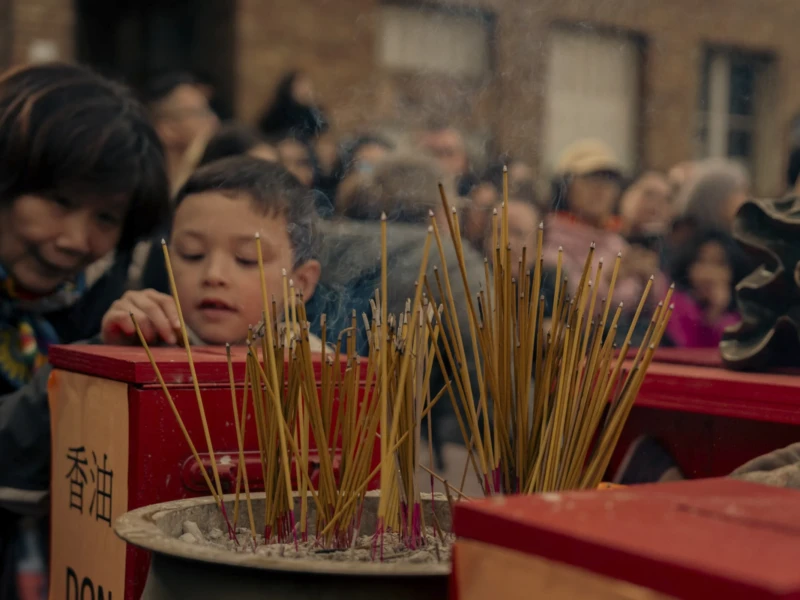 Cette photographie montre un enfant comme sujet principal, penché vers des bâtons d'encens allumés dans des boîtes rouges. La composition met en avant une scène religieuse ou culturelle, avec plusieurs personnes en arrière-plan.Le cadrage est serré sur l'enfant et les encensoirs, créant une ambiance intime. La lumière est douce et tamisée, évoquant un intérieur ou un lieu sacré. Les couleurs dominantes sont les tons chauds (rouge, or) et les teintes neutres, renforçant une atmosphère solennelle et spirituelle. Le style est documentaire et naturel, captant un moment authentique. L'ambiance est calme et respectueuse.