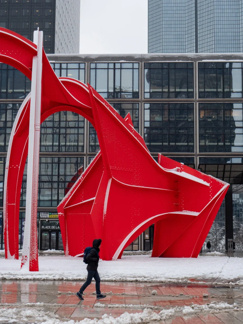 Daniel · 3888 x 5184 Cette photographie met en scène une grande sculpture rouge abstraite et architecturale, évoquant des formes dynamiques et organiques. Le sujet principal est cette installation, contrastant avec l'arrière-plan d'un bâtiment moderne en verre.Le cadrage est serré sur la sculpture, avec une personne en mouvement en premier plan, ajoutant une échelle humaine. La lumière semble naturelle, probablement hivernale, avec des tons froids et une ambiance grise et neigeuse. Les couleurs dominantes sont le rouge vif de la sculpture et les gris froids de la neige et du bâtiment. L'ambiance est à la fois moderne et contemplative, mêlant art urbain et nature hivernale.