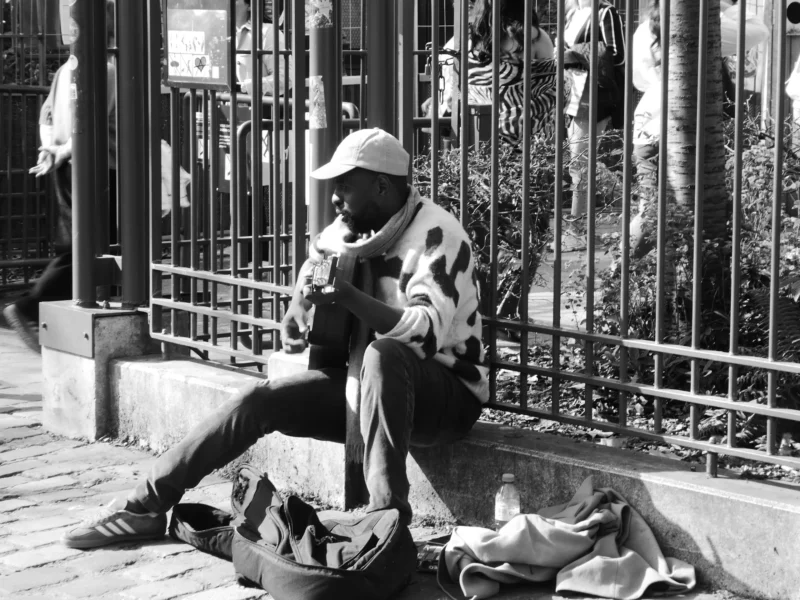 Christelle · 21/03/2026 · 2560 x 1920 Cette photographie en noir et blanc montre un homme assis comme sujet principal, absorbé par son téléphone portable. Il est vêtu d’un manteau et d’un chapeau, avec des bagages posés à côté de lui.La composition est simple et naturelle, avec un cadrage serré sur le sujet. L’arrière-plan, une grille métallique et des bâtiments, suggère un espace urbain. La lumière est douce, créant des contrastes modérés et une ambiance contemplative. Le style est documentaire, réaliste et dépouillé. L’ambiance est calme et introspective, évoquant la solitude en milieu urbain.