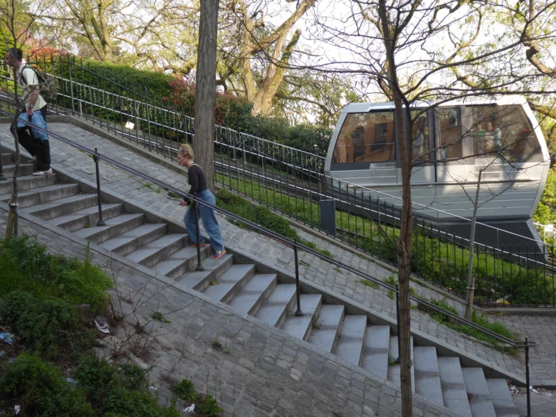 Christelle · 21/03/2026 · 4000 x 3000 Cette photographie montre une personne descendant un escalier extérieur en pierre menant à un funiculaire. Le sujet principal est cette personne, vêtue d’un jean et d’un haut sombre.La composition est dynamique, avec un cadrage légèrement en contre-plongée. Les lignes des marches et de la rampe guident le regard vers le funiculaire. La lumière est naturelle, douce et légèrement tamisée, créant des ombres douces. Les couleurs dominantes sont les tons verts (végétation), gris (pierres et métal) et quelques touches de bleu et de bois.L’ambiance est calme et paisible, évoquant une scène urbaine tranquille.