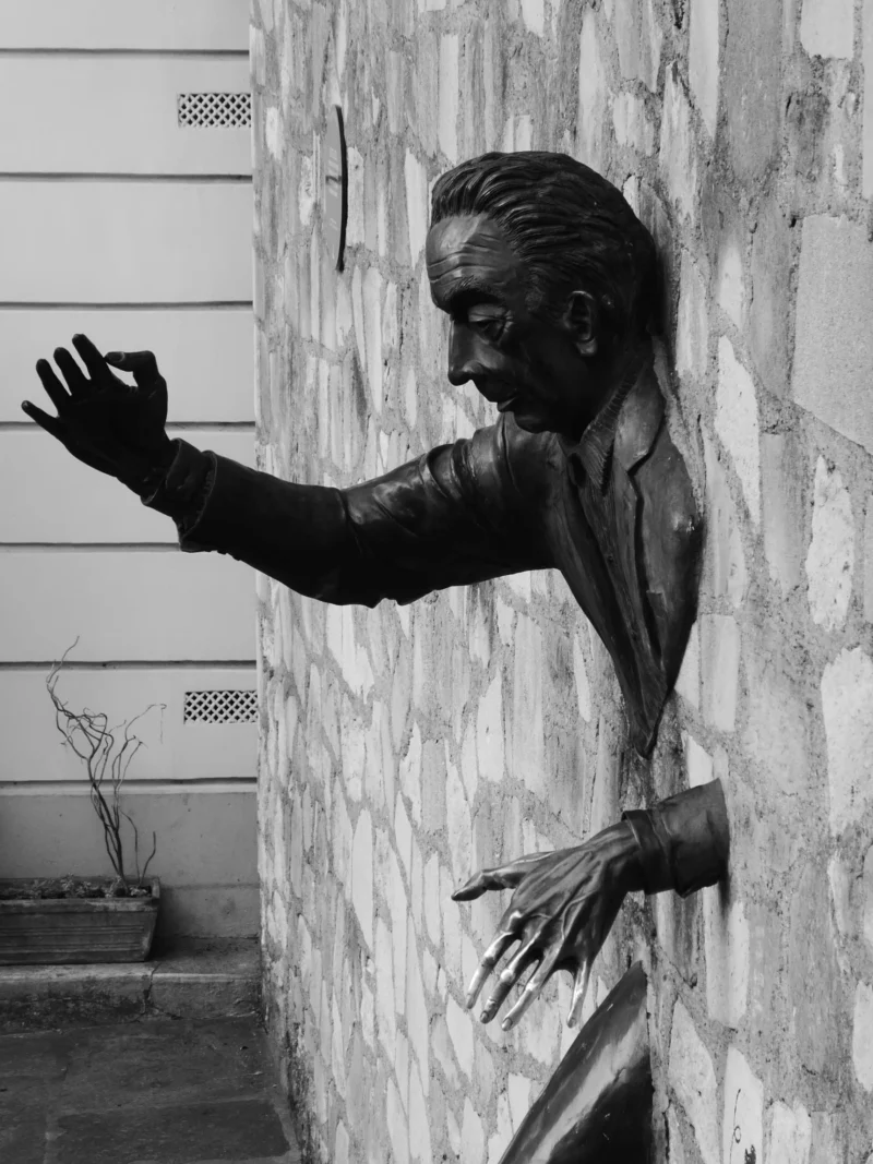 Christelle · 21/03/2026 · 2560 x 1920 Cette photographie montre une statue en bronze du sujet principal, un homme aux traits stylisés, en posture dynamique avec un bras tendu. La composition est simple, centrée sur la statue devant un mur blanc et une plante en pot. Le cadrage est serré, mettant en valeur les détails du visage et des mains.La lumière, naturelle et douce, crée des contrastes entre ombres et zones éclairées. Les couleurs dominantes sont les tons gris-brun de la statue et le blanc du mur, avec quelques touches de vert. Le style est réaliste mais épuré, évoquant une ambiance à la fois contemplative et puissante.