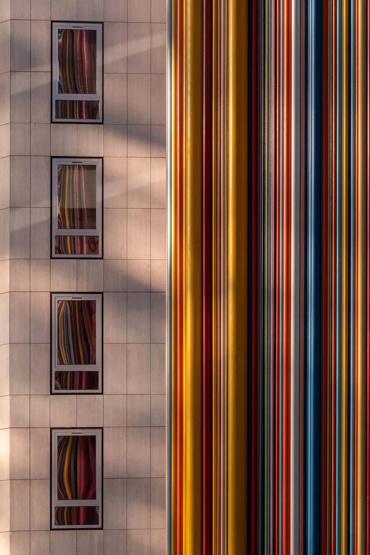 Daniel · 21/01/2019 · 3149 x 4724 Cette photographie met en scène un bâtiment moderne dont la façade est partiellement réfléchie sur une surface striée de couleurs verticales. Le sujet principal est cette superposition de motifs géométriques colorés (rouges, oranges, jaunes) avec les fenêtres rectangulaires du bâtiment.La composition joue sur les contrastes entre lignes droites et reflets. Le cadrage serré accentue les détails architecturaux et les jeux de lumière. Les couleurs dominantes sont chaudes et saturées, créant une ambiance vibrante et dynamique. Le style est épuré et contemporain, évoquant une esthétique urbaine et artistique.