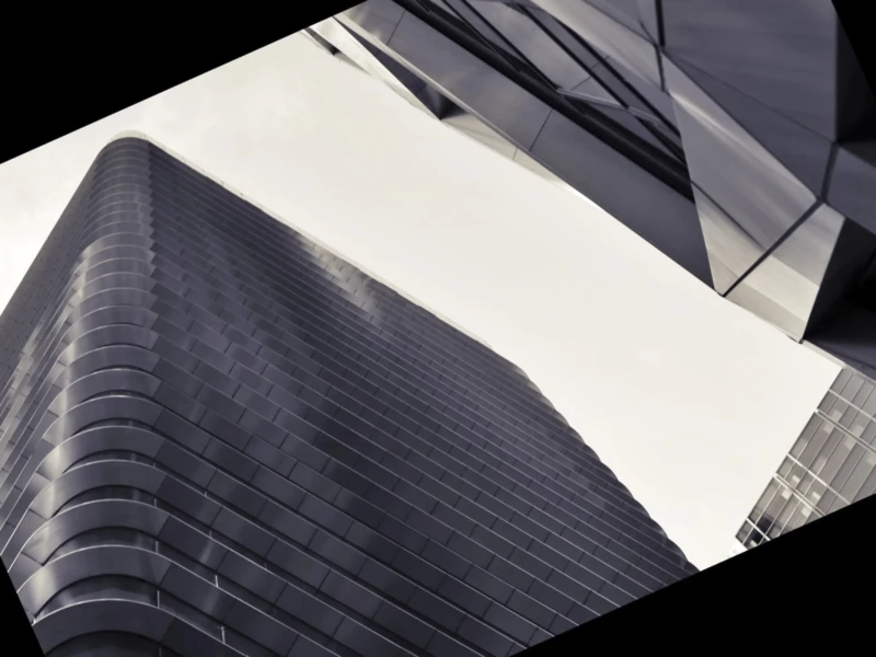 Cette photographie met en scène un **gratte-ciel moderne** aux lignes géométriques marquées, reflétant une **architecture contemporaine**. Le **cadrage serré et angulaire** accentue les angles aigus et les jeux de lumière.La **composition** joue sur les contrastes entre ombres et reflets, créant une ambiance **minérale et futuriste**. Les **couleurs dominantes** sont des tons **gris métallisés et blancs**, renforçant l’aspect froid et high-tech.Le **style** est épuré, presque abstrait, avec une **lumière rasante** qui souligne les textures et les arêtes. L’ambiance est **urbaine, dynamique et légèrement mystérieuse**.