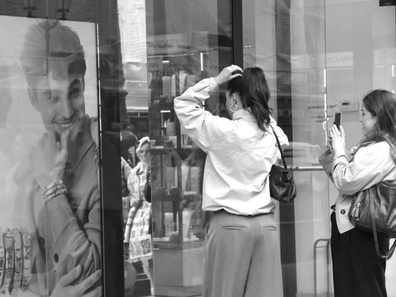 Cette photographie en noir et blanc montre deux femmes observant une œuvre d’art abstraite ou pixelisée représentant un visage masculin. Le sujet principal est cette toile, encadrée et accrochée au mur.La composition est dynamique, avec un cadrage serré sur les femmes et l’art. La lumière semble naturelle et douce, éclairant les visages et l’œuvre. L’ambiance est contemplative, presque intimiste, renforcée par les tons de gris et les jeux d’ombres. Le style est épuré, mettant l’accent sur les expressions et les interactions des personnages.