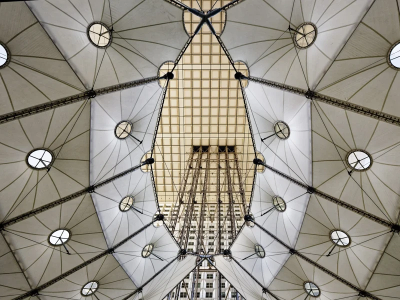 Cette photographie montre une **structure architecturale complexe**, probablement une verrière ou un dôme géodésique. Le **sujet principal** est la **charpente métallique** et les **lucarnes circulaires** qui laissent filtrer la lumière.La **composition** est symétrique et géométrique, avec des lignes convergentes vers le centre. Le **cadrage** est serré, mettant en valeur les détails techniques. La **lumière**, naturelle et tamisée, crée des contrastes doux. Les **couleurs dominantes** sont des tons neutres (gris, blanc, beige), renforçant une ambiance **minérale et industrielle**.Le style évoque une **architecture moderne et ingénierie sophistiquée**, avec une ambiance **austère et futuriste**.