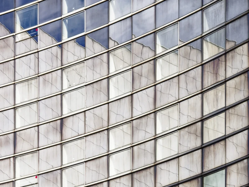 Cette photographie met en scène une façade moderne de bâtiment composée de nombreux panneaux de verre et de métal. Le sujet principal est l’architecture géométrique et répétitive des fenêtres rectangulaires.Le cadrage est serré, centré sur les éléments structuraux, créant une composition symétrique et rigoureuse. La lumière, naturelle et diffuse, accentue les reflets et les nuances des matériaux. Les couleurs dominantes sont des tons gris et blancs, renforçant une ambiance minimaliste et contemporaine. Le style épuré et industriel évoque une esthétique urbaine et sophistiquée, évoquant modernité et précision architecturale.