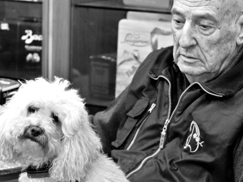 Cette photographie met en scène un homme âgé assis, tenant un petit chien blanc à poils longs. Le sujet principal est ce duo, créant une ambiance chaleureuse et intime.La composition est simple et centrée, avec un cadrage serré sur les deux personnages. L’arrière-plan, légèrement flou, met l’accent sur eux. La lumière douce et naturelle, probablement en noir et blanc, accentue les contrastes et les textures, comme les rides du visage et le pelage du chien. Le style est documentaire et réaliste, évoquant une scène du quotidien. L’ambiance est à la fois douce et mélancolique.