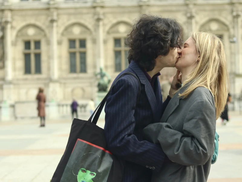 Cette photographie montre deux jeunes qui s’embrassent tendrement en extérieur. Le **sujet principal** est ce couple, au premier plan, avec une **composition** centrée sur eux.Le **cadrage** est serré, mettant en valeur leur intimité. L’arrière-plan, flou, révèle un bâtiment classique et des passants, suggérant une ville européenne. La **lumière** semble naturelle, douce et légèrement tamisée, créant une ambiance chaleureuse.Les **couleurs dominantes** sont sobres : gris, noir et tons pastel, renforçant une atmosphère romantique et nostalgique. Le style évoque un **cinéma réaliste**, avec une touche mélancolique et poétique.
