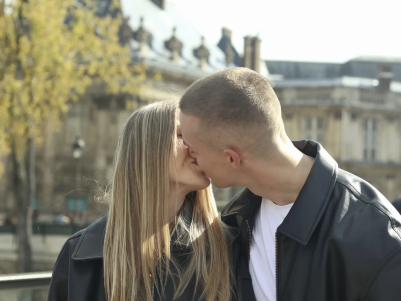 Cette photographie met en scène un couple en train de s'embrasser tendrement. Le sujet principal est centré sur leurs visages, avec un cadrage serré et une composition naturelle.La lumière est douce et naturelle, probablement en journée, éclairant leurs traits avec une ambiance chaleureuse. Les couleurs dominantes sont des tons neutres (noir, gris) avec des touches de vert et de beige en arrière-plan.Le style est simple et spontané, évoquant une scène intime et romantique. L’ambiance est douce et émouvante, renforcée par le flou de l’arrière-plan.