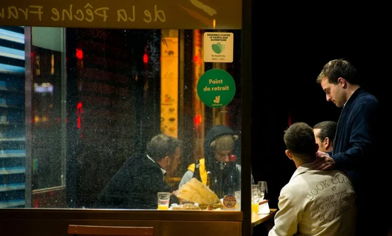 Pascale · 09/11/2025 · 1000 x 604 Cette photographie capture l'intérieur d'un restaurant ou d'une boulangerie la nuit. Le sujet principal semble être un groupe de personnes assises à une table, éclairées par une lumière chaude et tamisée.La composition montre un cadrage serré sur les clients, avec une vitrine reflétant des enseignes lumineuses à l'extérieur. Les couleurs dominantes sont des tons chauds (jaunes, oranges) contrastant avec les teintes sombres de la nuit.Le style est réaliste et spontané, évoquant une ambiance conviviale et intimiste, presque nocturne et légèrement animée.