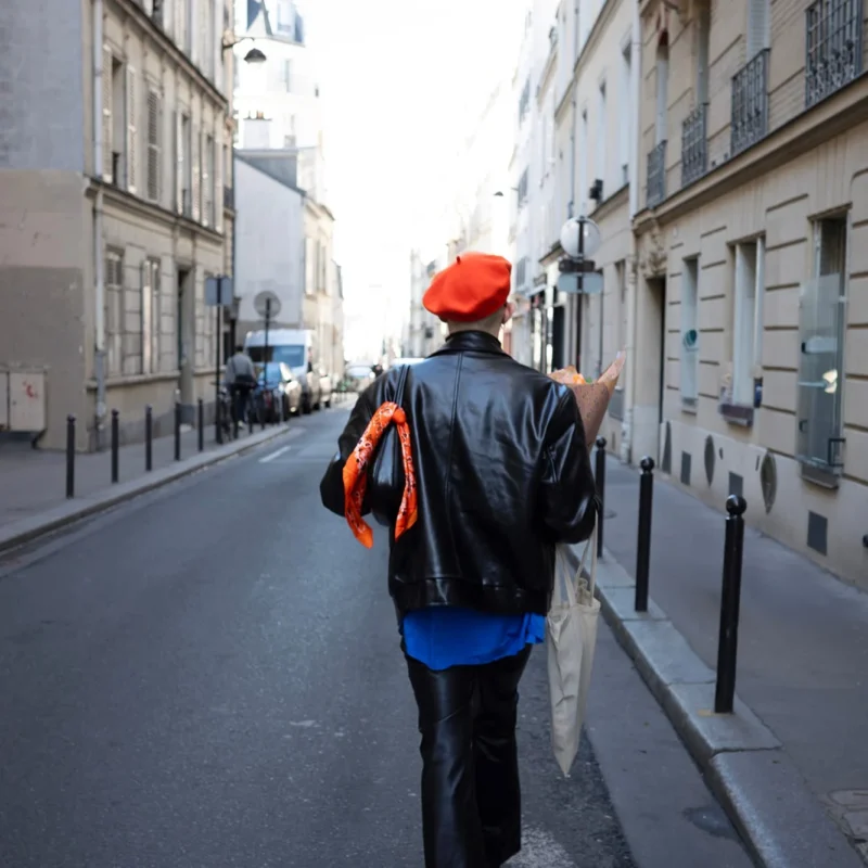 Nathalie C H · 1333 x 1333 Cette photographie montre une personne vue de dos, marchant dans une rue étroite et pavée. Le sujet principal porte un manteau noir, un béret rouge et un sac à dos orange.La composition est centrée sur la silhouette, avec une perspective légèrement basse. Le cadrage est serré, mettant en valeur les détails de la rue et des bâtiments.La lumière est naturelle, douce et légèrement voilée, créant une ambiance calme et un peu mélancolique. Les couleurs dominantes sont les tons neutres des bâtiments, rehaussés par les touches de rouge et orange du sujet. Le style est documentaire et réaliste.