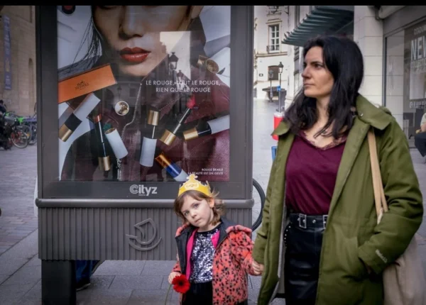 Muriel Juan · 1080 x 774 Cette photographie montre une femme et un enfant comme sujets principaux. La composition met en valeur leur interaction devant une affiche publicitaire colorée. Le cadrage est serré sur eux, avec l’affiche en arrière-plan.La lumière semble naturelle, avec des ombres douces. Les couleurs dominantes sont les tons froids (bleu, gris) et chauds (marron, rouge), créant un contraste. Le style est réaliste et documentaire, capturant un moment du quotidien. L’ambiance est à la fois urbaine et intime, évoquant une promenade en ville.