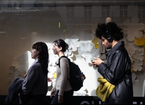Muriel Juan · 1080 x 778 Cette photographie capture trois jeunes en intérieur, probablement dans un lieu public comme un café ou un espace culturel. Le sujet principal semble être le trio, avec une attention particulière sur la personne à droite, qui regarde son téléphone.La composition est dynamique, avec un cadrage serré sur les visages et les gestes. La lumière est tamisée, créant une ambiance intimiste et légèrement dramatique. Les couleurs dominantes sont des tons neutres et sombres, avec des éclairages chauds qui contrastent avec les teintes froides des murs.Le style est réaliste et documentaire, évoquant une ambiance moderne et urbaine.