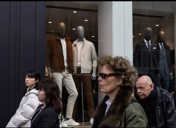Muriel Juan · 1080 x 783 Cette photographie montre une vitrine de boutique de vêtements masculins. Le sujet principal est une mannequin vêtu de costumes, reflété par les vitres.La composition met en scène des passants observant la vitrine, créant une ambiance urbaine. Le cadrage est serré sur les visages et les vêtements, avec une profondeur limitée.La lumière semble naturelle, bien que légèrement tamisée, typique d’un jour d’hiver. Les couleurs dominantes sont neutres : gris, beige et noir, renforçant l’aspect élégant et sobre.Le style est documentaire, capturant une scène de vie quotidienne dans un cadre urbain. L’ambiance est à la fois dynamique et calme.