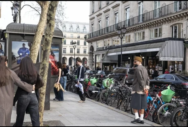 Muriel Juan · 1080 x 730 Cette photographie capture une scène urbaine animée. Le sujet principal est un groupe de personnes sur une rue piétonne, avec des vélos attachés en premier plan. La composition montre une perspective légèrement basse, mettant en valeur les passants et les bâtiments en arrière-plan.Le cadrage est serré, centré sur les interactions humaines. La lumière est naturelle, probablement en journée, avec des ombres douces. Les couleurs dominantes sont neutres et grises, adoucies par des touches de vert (arbres) et de bleu (vêtements). Le style est documentaire, réaliste, reflétant une ambiance quotidienne et vivante dans une ville européenne.