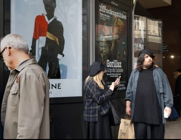 Muriel Juan · 1080 x 833 Cette photographie montre trois personnes en extérieur devant une affiche publicitaire pour le film *« Gorillaz »*.Le sujet principal semble être les deux femmes en premier plan, engagées dans une conversation, tandis que l’homme à gauche observe. La composition est dynamique, avec un cadrage serré sur les visages et les détails vestimentaires. La lumière est naturelle, légèrement tamisée, créant des contrastes doux. Les couleurs dominantes sont sobres : gris, noir, et touches de rouge et bleu sur l’affiche.Le style est réaliste et documentaire, capturant une scène urbaine authentique. L’ambiance est calme et quotidienne, avec une touche de mystère apportée par l’affiche cinématographique.