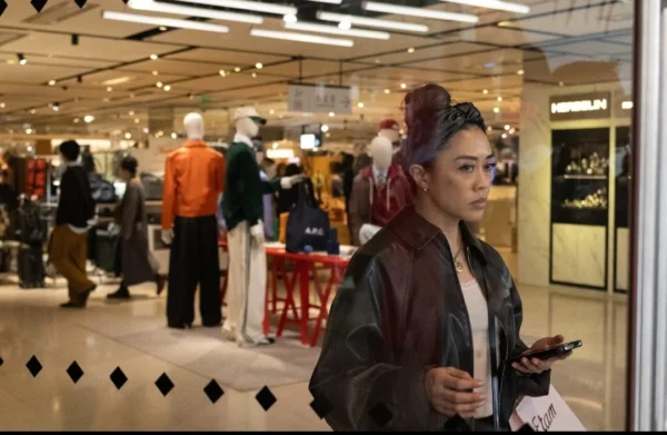 Muriel Juan · 1080 x 703 Cette photographie montre une jeune femme comme sujet principal, vêtue d’une veste longue et coiffée d’une perruque aux reflets violets, tenant un téléphone. L’arrière-plan révèle un magasin ou un espace commercial animé, avec des mannequins et des clients.Le cadrage est serré sur elle, mais inclut des éléments contextuels. La lumière est artificielle et uniforme, typique d’un éclairage intérieur. Les couleurs dominantes sont neutres (beige, gris) avec des touches vives comme les reflets violets de la perruque et les vêtements colorés des mannequins.L’ambiance est urbaine et moderne, avec une touche de fantaisie apportée par la perruque.