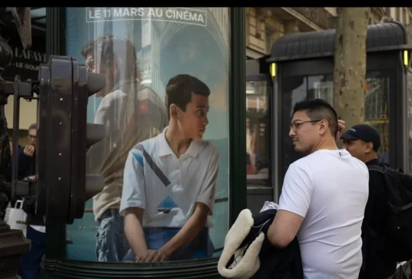 Muriel Juan · 1080 x 732 Cette photographie montre deux hommes dans un cadre urbain. Le sujet principal semble être le reflet d’un individu dans une vitrine, tandis que l’autre homme, à l’extérieur, observe la scène. La composition joue sur les reflets et les contrastes entre intérieur et extérieur. Le cadrage est serré, mettant en valeur les visages et les détails des vêtements.La lumière est naturelle, légèrement tamisée, créant des ombres douces. Les couleurs dominantes sont des tons neutres (gris, blanc) avec des touches de bleu et de noir. Le style est réaliste et documentaire, capturant un moment spontané. L’ambiance est à la fois urbaine et introspective, évoquant une réflexion sur la solitude en ville.