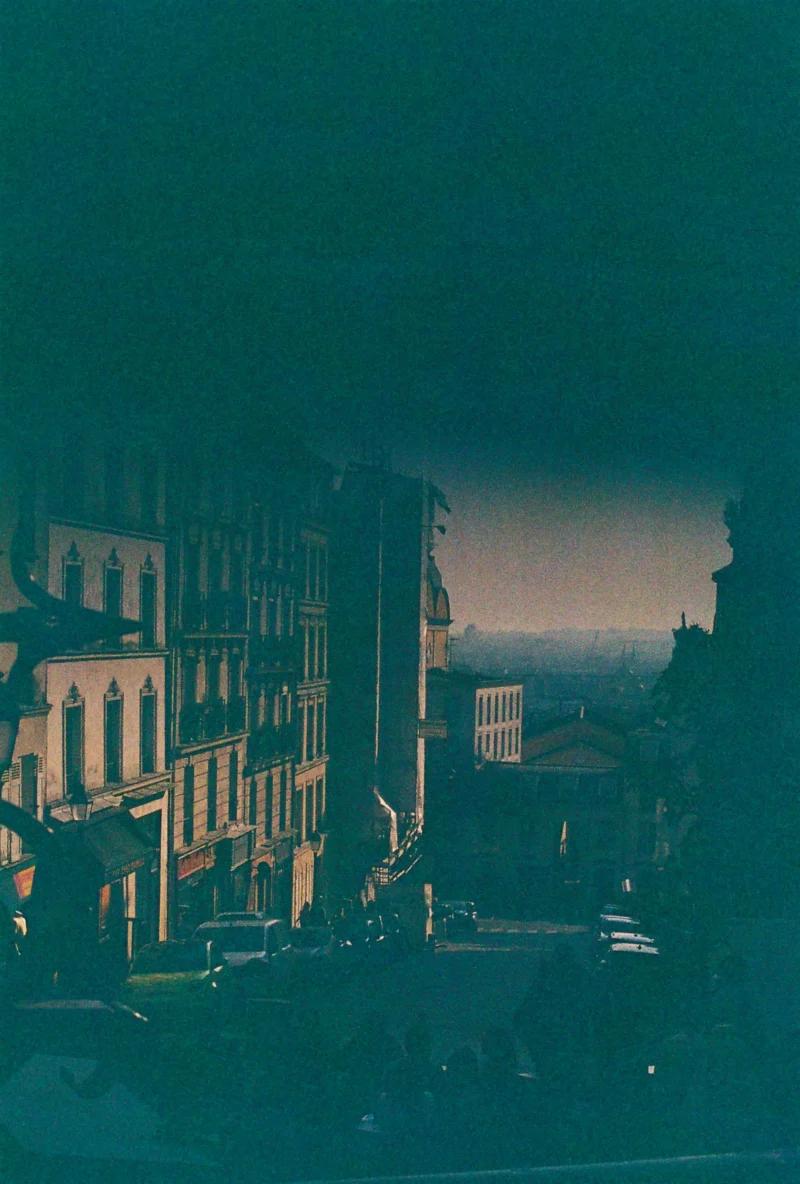 Nadia · 2433 x 3601 Cette photographie capture une ville en soirée avec une ambiance urbaine mélancolique. Le sujet principal semble être un ensemble d'immeubles anciens et une rue déserte.La composition est symétrique, avec des bâtiments alignés de chaque côté. Le cadrage est légèrement bas, offrant une perspective élargie vers le ciel. La lumière est douce et tamisée, créant des ombres longues et des teintes chaudes contrastant avec le bleu dominant.Les couleurs dominantes sont des tons bleus et bruns, avec des reflets dorés. Le style évoque une esthétique vintage, renforcée par les teintes sépia. L'ambiance est calme et nostalgique, presque onirique.
