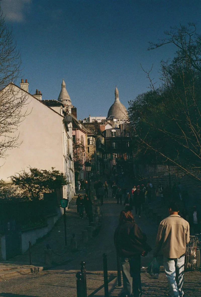 Nadia · 2433 x 3601 Cette photographie capture un groupe de personnes en extérieur, probablement dans un lieu touristique. Le sujet principal semble être l’architecture historique en arrière-plan, avec des bâtiments aux toits arrondis et une tour distinctive, évoquant un style médiéval ou gothique.La composition est dynamique, avec un cadrage incliné et des personnes en mouvement. La lumière est naturelle, douce et légèrement dorée, suggérant un moment en fin de journée. Les couleurs dominantes sont des tons neutres (beige, gris) avec des touches de vert des arbres et de bleu du ciel.L’ambiance est à la fois animée et paisible, mêlant vie quotidienne et patrimoine architectural.