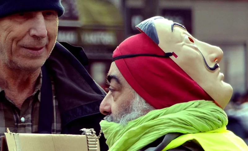 Pascale · 11/01/2020 · 1000 x 610 Cette photographie met en scène deux hommes comme sujets principaux. L’un porte un masque coloré en forme de visage, tandis que l’autre semble l’observer. La composition est serrée, centrée sur leurs visages.Le cadrage est rapproché, mettant l’accent sur les expressions et les détails des accessoires. La lumière semble naturelle, douce et légèrement tamisée, créant des ombres douces.Les couleurs dominantes sont vives : rouge du masque, vert du foulard, et tons neutres des vêtements. Le style est documentaire et spontané, évoquant une ambiance à la fois humoristique et mystérieuse.